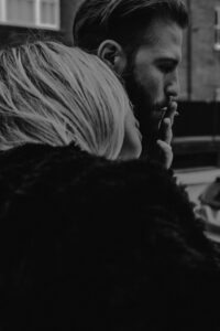 A close-up black and white photo of a couple intimately sharing a smoke outdoors.