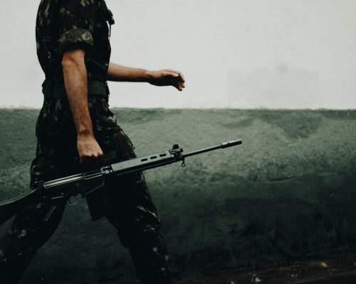 A soldier in camouflage walking with a rifle against a textured wall, day.